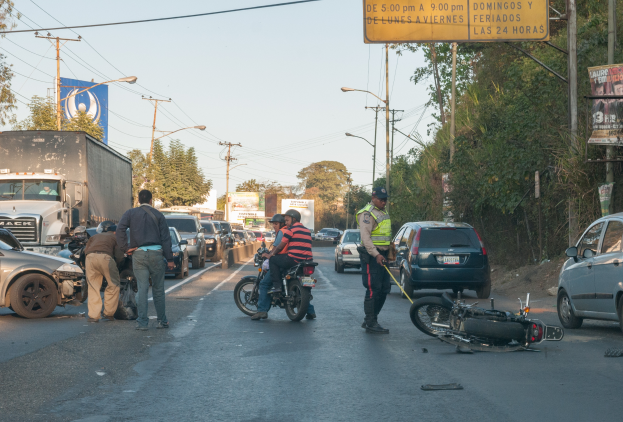 Eine Gruppe von Menschen um ein verunglücktes Motorrad auf der Straße herumsteht, umgeben von mehreren Fahrzeugen, einschließlich eines Lastwagens, und einer Hintergrundlandschaft aus Bäumen, Pfählen, Lampen, Schildern und Himmel.