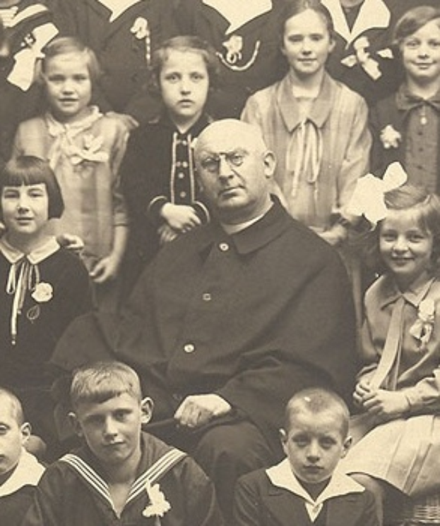 Ein altes Schwarz-Wei├č-Foto eines ernst dreinschauenden Mannes, der in der Mitte sitzt und von einer Gruppe Kinder umgeben ist, die alle bewundernd zu ihm aufschauen.