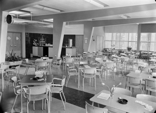 Schwarzes und weißes Foto einer Cafeteria mit Tischen und Stühlen, Tellern, Besteck, Gläsern und Blumenvasen auf den Tischen und einer Wand mit Türen, Fenstern und Deckenleuchten im Hintergrund.