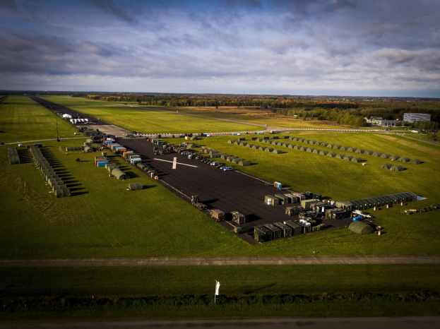 Luftaufnahme einer Militärbasis in den Niederlanden mit Straßen, Gras, Bäumen, Gebäuden, Fahrzeugen und einem bewölkten Himmel.