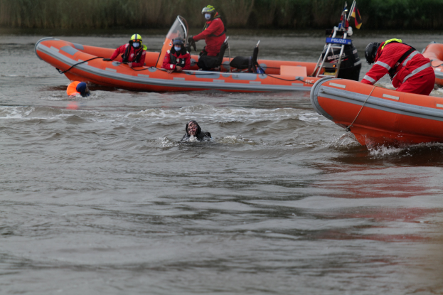Gruppe von Menschen in einem Schlauchboot auf einem Fluss, mit zwei Personen im Wasser im Vordergrund und Vegetation im Hintergrund, alle tragen Schwimmwesten und Helme bei einer Rettungsaktion.