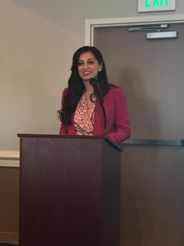 Eine Frau steht an einem Rednerpult mit einem Mikrofon, lächelt und hält eine Rede in einem Raum mit einem Schild darüber, das 'Dr. Shabana Aziz' lautet.