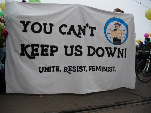 Eine Gruppe von Menschen steht auf einer Straße und hält ein Banner in der Hand, auf dem "You Can't Keep Us Down, Unite Resist Feminist" steht, mit einem Fahrrad in der Nähe und Ballons, Drähten und einem klaren blauen Himmel im Hintergrund.