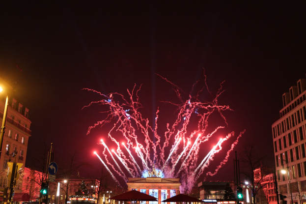 Eine belebte Stadtstraße an Silvester in Berlin, voller Menschen, Fahrzeuge und erleuchtet von Gebäudelichtern und Feuerwerk am Himmel.