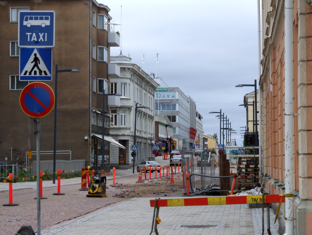 Stadtstraße mit Gebäuden, Fahrzeugen, Verkehrszeichen, Baustelle, Bäumen und Wolkenhimmel.