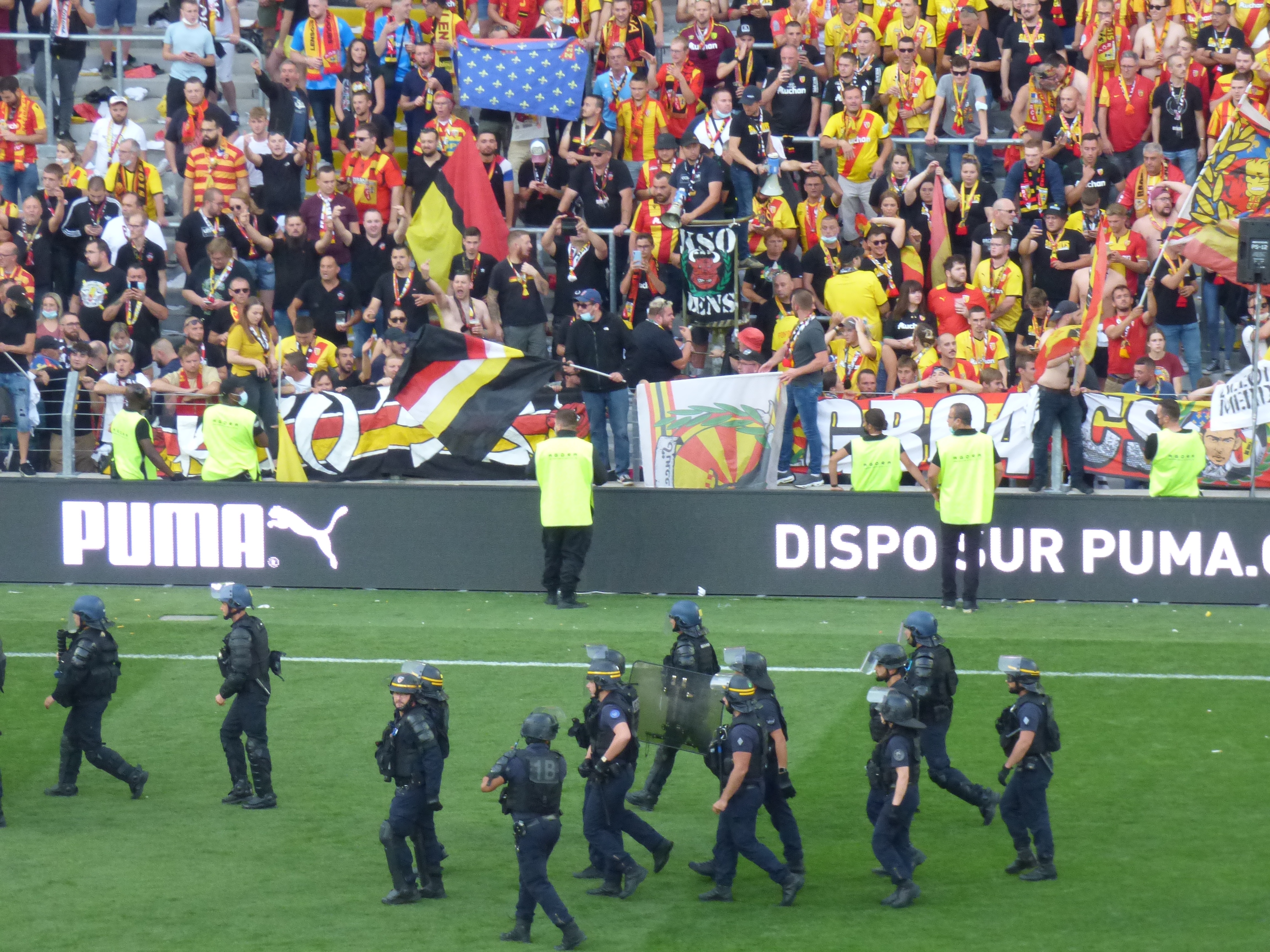 Eine Gruppe von Polizisten in Helmen und mit Waffen in der Hand läuft über ein Fußballfeld, im Hintergrund halten Menschen Fahnen und Schilder.