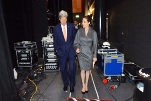 Angelina Jolie und John Kerry auf einem Boden umgeben von Menschen, Geräten, Kabeln, einem Helm auf einem Wagen, einem Müllbehälter, einem Vorhang, einer Säule und einem Dach mit Deckenleuchten.