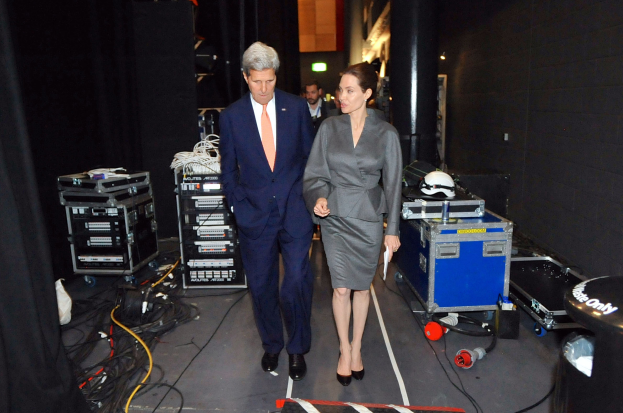 Angelina Jolie und John Kerry auf einem Boden umgeben von Menschen, Geräten, Kabeln, einem Helm auf einem Wagen, einem Müllbehälter, einem Vorhang, einer Säule und einem Dach mit Deckenleuchten.
