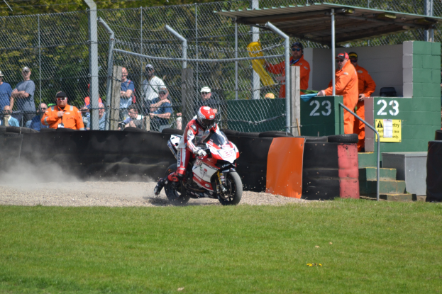Eine Person in Helm und Handschuhen fährt ein Motorrad über ein grasbewachsenes Feld, mit Zuschauern in Kappen und Schutzbrillen, Maschendrahtzaun, Treppe mit Geländer und einem nummerierten Schuppen im Hintergrund.
