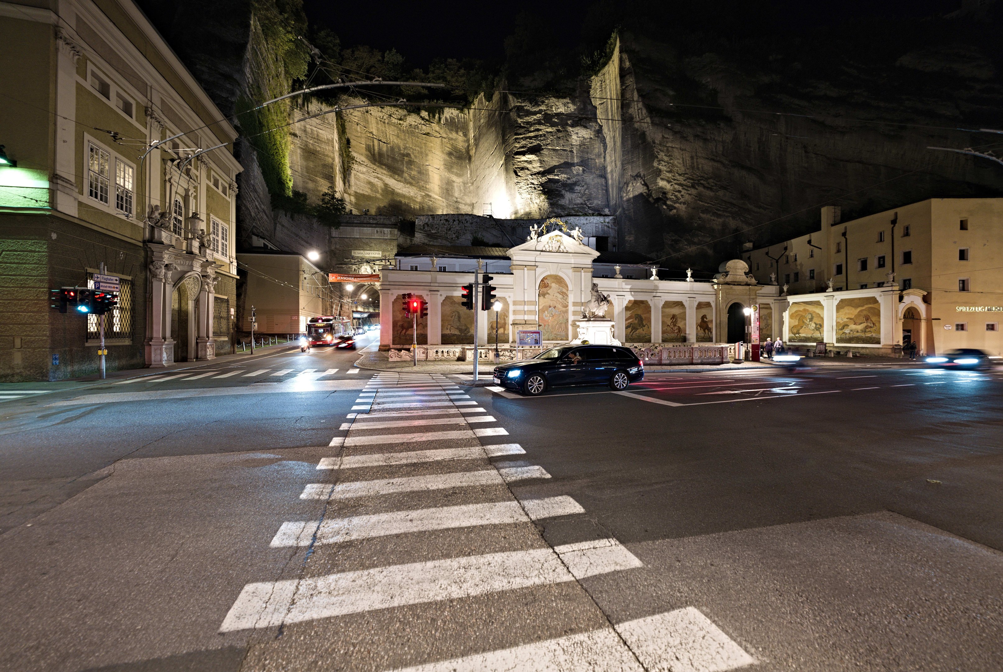Eine nächtliche Stadtstraße mit fahrenden Autos, Straßenlaternen, Ampeln, beleuchteten Fenstern in Gebäuden, Laternenpfählen, einem Hügel im Hintergrund und einer Zebrastreifenmarkierung am Boden.
