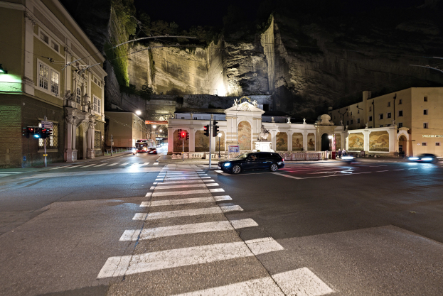 Eine nächtliche Stadtstraße mit fahrenden Autos, Straßenlaternen, Ampeln, beleuchteten Fenstern in Gebäuden, Laternenpfählen, einem Hügel im Hintergrund und einer Zebrastreifenmarkierung am Boden.