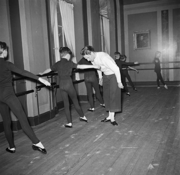 Eine Gruppe junger Tänzer in einem Tänzstudio, die vor einer Wand mit einem gerahmten Schwarz-Weiß-Foto eines Choreografen stehen, mit Fenstern und Vorhängen im Hintergrund.