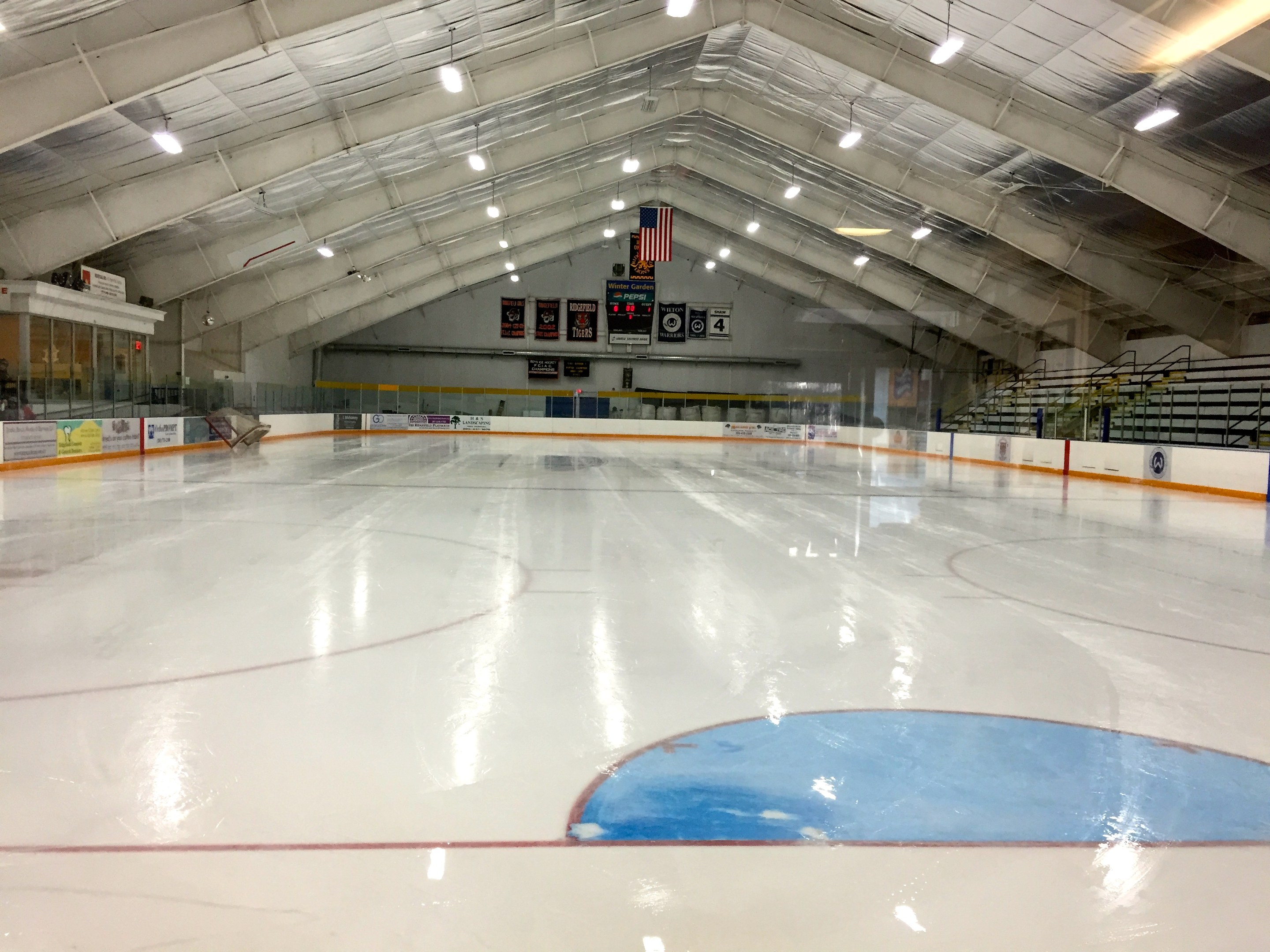 Große Indoor-Eisbahn mit Deckenbeleuchtung, Werbetafeln, einer Rückwand, Treppen mit Geländern und einigen Menschen, was auf eine Sportanlage hindeutet, wahrscheinlich eine Hockeyarena.