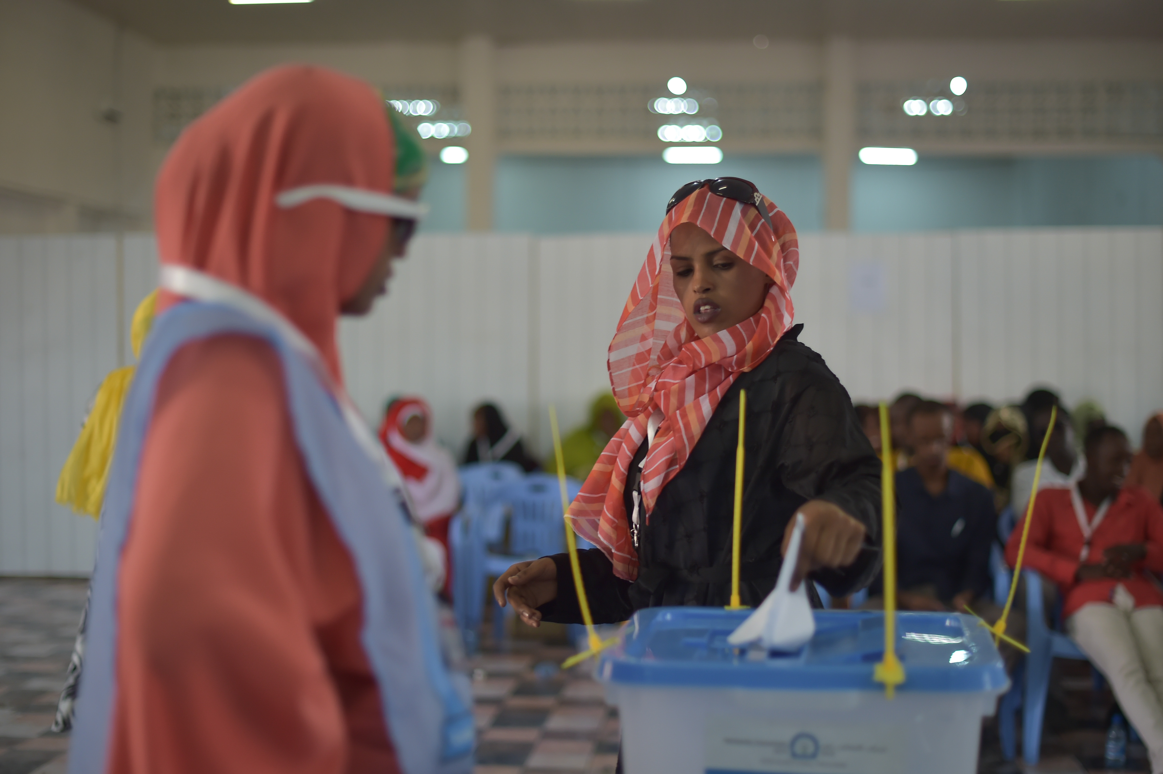 Eine Frau mit Kopftuch stimmt an einer Wahlurne ab, mit Menschen im Hintergrund und einer Box im Vordergrund, unter einer Decke mit Lampen und einer Wand dahinter.