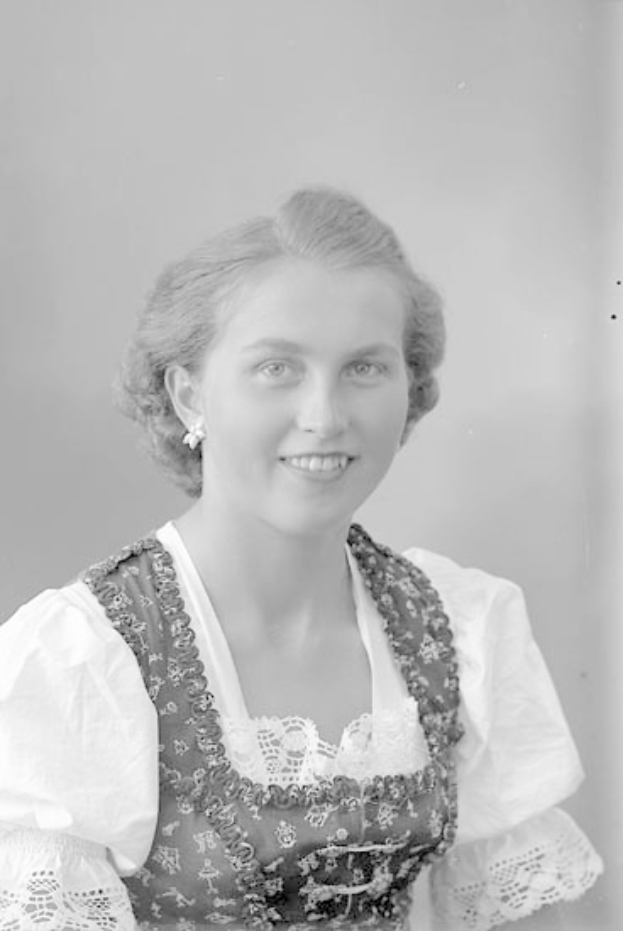 Ein Schwarz-Weiß-Foto einer Frau in traditioneller bayrischer Tracht, die herzlich lächelt und ihr Haar zu einem Dutt gebunden hat.