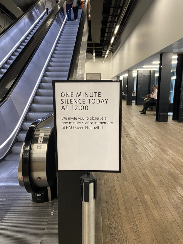 Eine Rolltreppe im Flughafen mit einem Schild, auf dem "Eine Minute Stille heute" steht, ein paar Menschen darauf und Lampen an der Decke im Hintergrund.