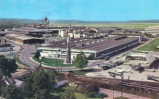 Altes Schwarz-Weiß-Foto eines Flughafens mit parkenden Flugzeugen, Gebäuden, Bäumen, Straßen, Fahrzeugen, Strommasten, Gras, Bahnschienen, Türmen, Hügeln und einem klaren blauen Himmel, mit Text unten.