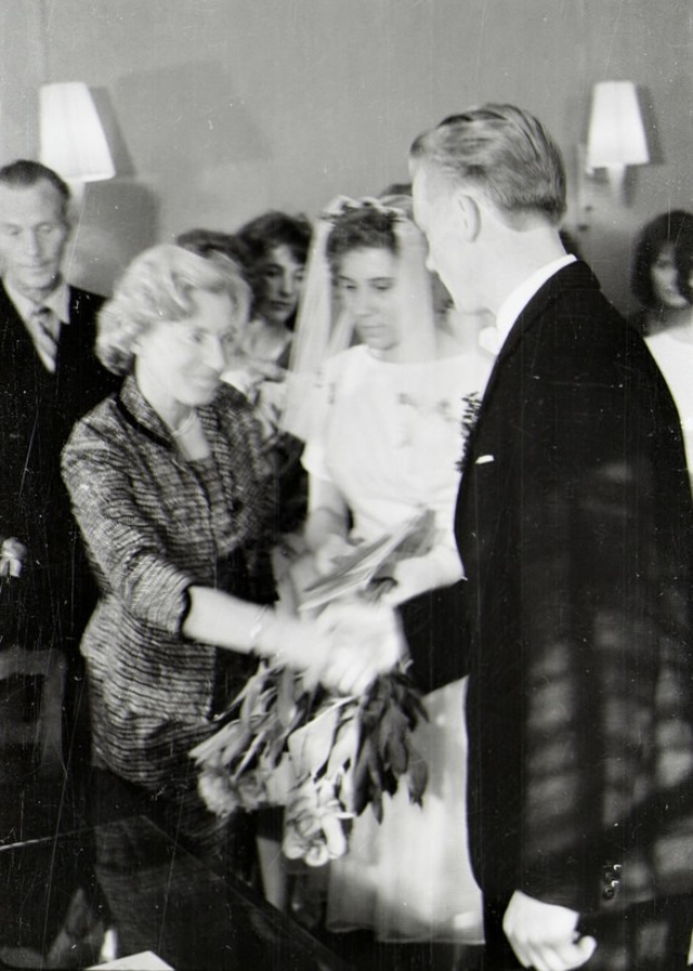 Ein Schwarz-Weiß-Foto einer Braut und eines Bräutigams beim Austausch der Eheringe, wobei die Braut ein weißes Kleid und einen Schleier trägt und einen Strauß hält, und der Bräutigam einen Anzug und eine Krawatte trägt, mit Wandlampen im Hintergrund.