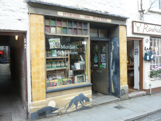 Ein Geschäft namens 'Mord und Totschlag' mit Regalen voller Bücher, verschiedener Gegenstände, Texttafeln und Pflanzen mit Blumen, mit einem Baum und einer Straße im Vordergrund.