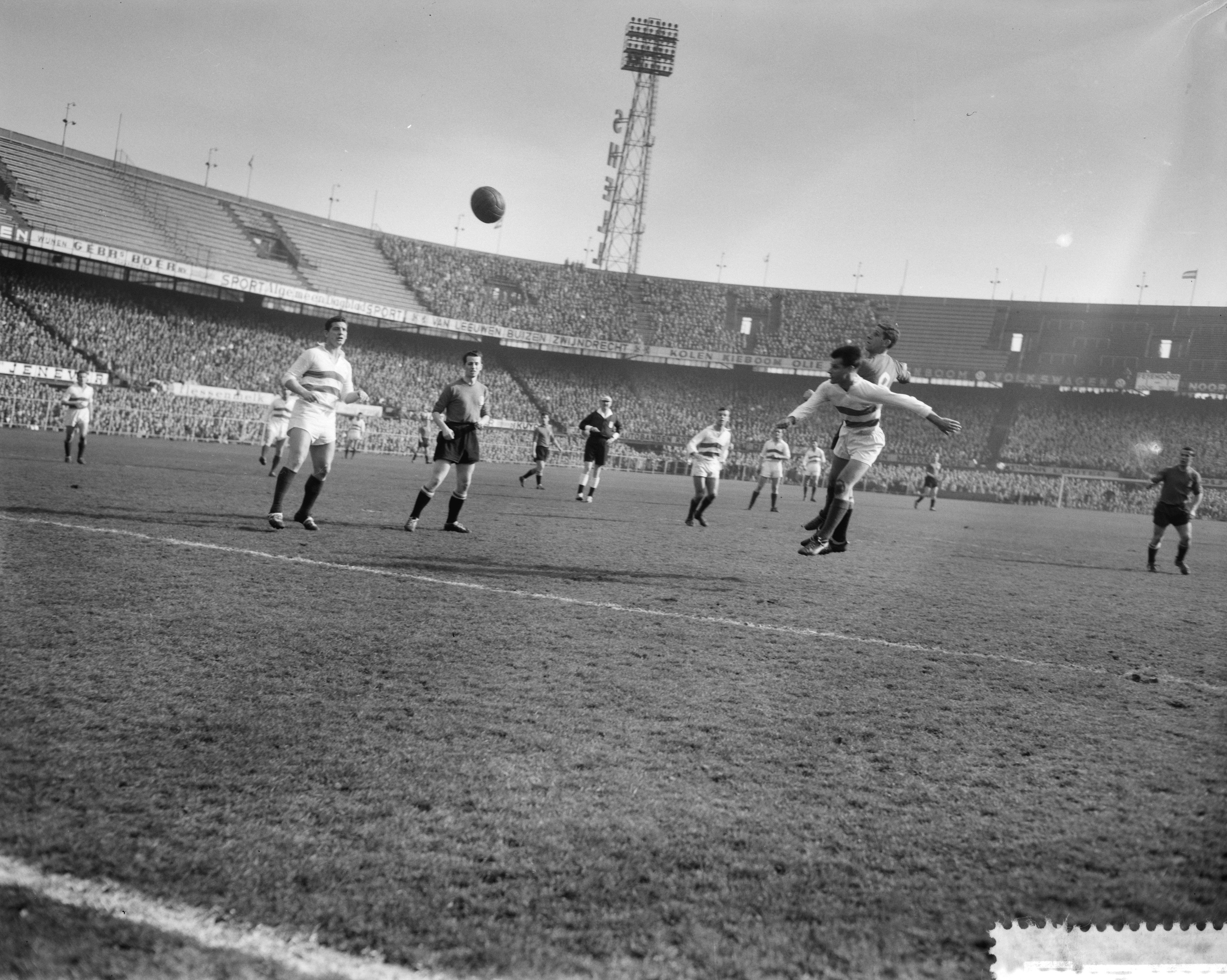 Eine Gruppe von Männern, die auf einem Feld mit einem Stadion, Flutlicht und einem klaren Himmel im Hintergrund Fußball spielen.