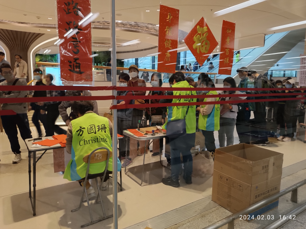 Eine Gruppe von Menschen mit Mund-Nasen-Schutz steht an einem Tresen in einem Einkaufszentrum. Es sind Stühle und Tische mit verschiedenen Gegenständen darauf zu sehen, und eine Person sitzt auf einem der Stühle. Vor dem Tresen befindet sich ein Geländer, und von der Decke hängen Schilder mit Text. Rechts im Bild befindet sich eine Kartonschachtel und ein Wasserzeichen.