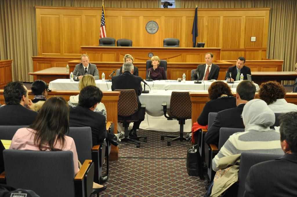 Ein Versammlungsraum mit einer Gruppe von Menschen, die auf Stühlen sitzen, zwei √§ußere Flaggen, ein oranger Schreibtisch mit Wasserflaschen und einem Mikrofon.