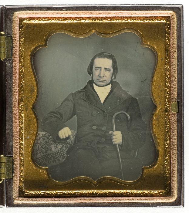 Ein altes Daguerreotyp eines Mannes in einem Anzug, der einen Stock hält und ernst in die Ferne blickt.