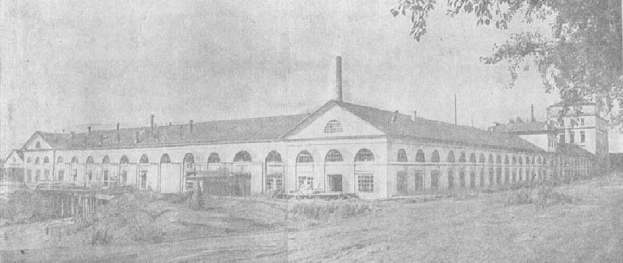 Schwarz-weiß-Foto eines großen, mehrfenstrigen Gebäudes, das von Bäumen und Pflanzen umgeben ist und unter einem klaren Himmel steht, mit dem Text "1892 – das erste Fabrikgebäude der Stadt" unten.