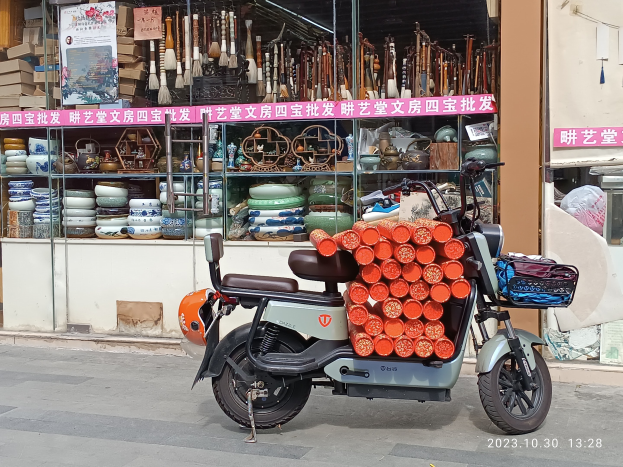 Ein Motorroller mit einem Korb steht auf der Straße vor einem Laden mit einer Glaswand, auf der verschiedene Gegenstände wie Schalen und Teller zu sehen sind, sowie eine Tafel mit Text im Inneren.
