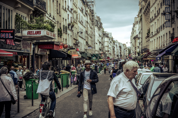 Zwei große Gebäude mit einer Straße dazwischen, auf der eine Menschenmenge geht und mehrere Stände im Erdgeschoss aufgestellt sind; ein Mann fährt mit dem Fahrrad auf der Straße, und einige Autos sind auf der gegenüberliegenden Seite geparkt.