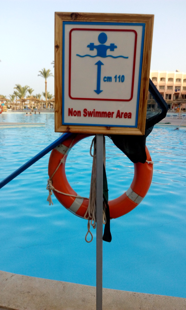 Ein Schwimmbad mit einem Schild "Nur für Nichtschwimmer" und einem Rettungsring an einem Pfahl, umgeben von Bäumen, Gebäuden und einem klaren blauen Himmel.