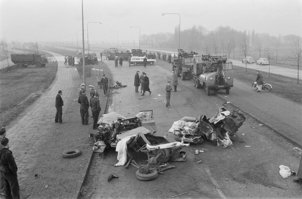 Schwarz-weiß-Szene eines Autounfalls am Straßenrand mit mehreren Fahrzeugen, Einsatzkräften und Zuschauern, Laternenpfähle, Bäume und Himmel im Hintergrund.