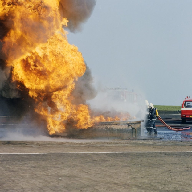 Ein Feuerwehrauto steht in Flammen an der Seite einer Straße, mit zwei Personen in Helmen in der Nähe, die Rohre halten, und einem Fahrzeug und dem Himmel im Hintergrund.