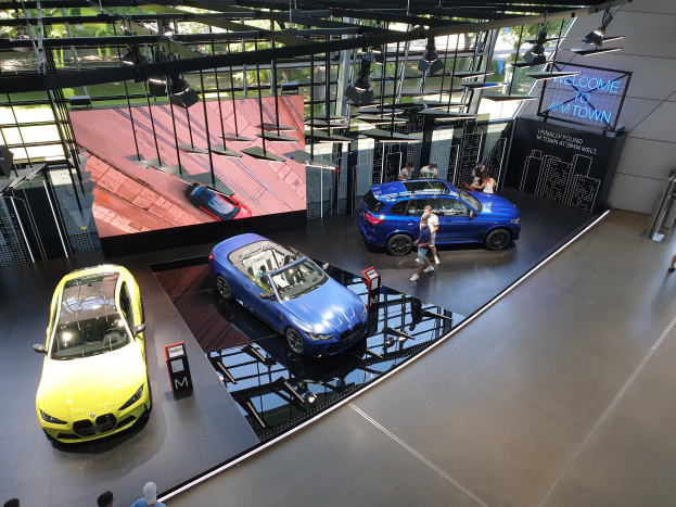 Eine Gruppe von Menschen steht im BMW Museum in München, Deutschland, mit ausgestellt Autos auf der linken Seite und einem Schild mit der Aufschrift "BMW Museum München" auf der rechten Seite, das auf eine Außenansicht von Bäumen, Gebäuden und Himmel durch Glasfenster blickt.