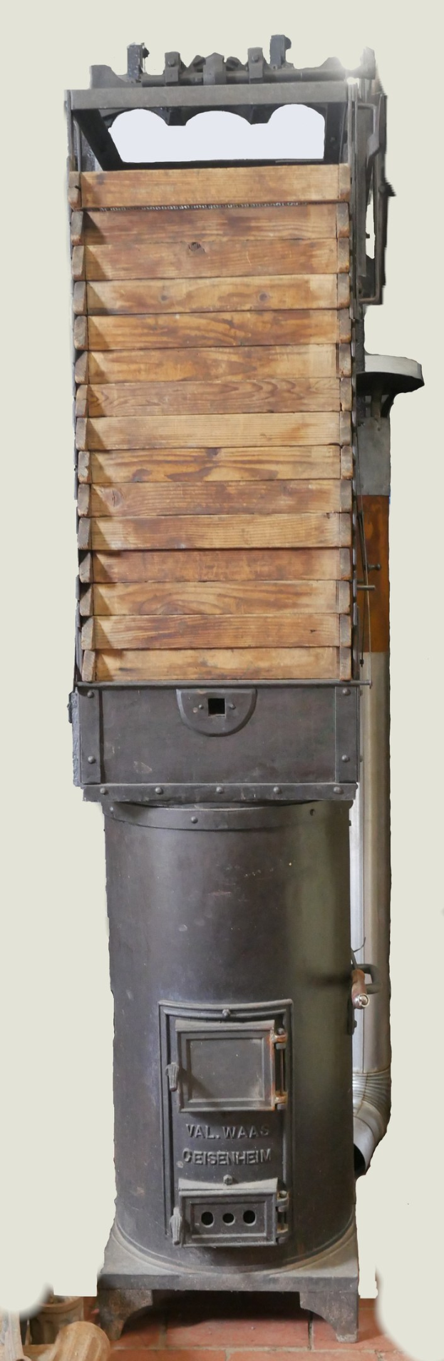 Altmodischer Holzofen mit Metall- und Holzkonstruktion, Schornstein, auf einem Ziegelboden mit einer Wand im Hintergrund.