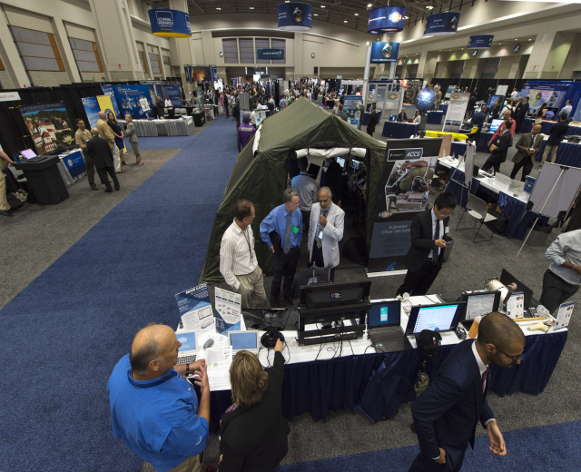 Große Gruppe von Menschen auf einer Karriere-Messe um Tische mit Laptops, Monitoren und Papieren unter einem Zelt mit Schildern, Deckenleuchten und Fenstern im Hintergrund.