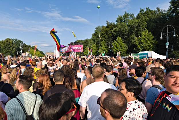 Eine große Menschenmenge steht vor einem Gebäude, einige halten Fahnen und Banner, mit einem Zelt, Laternenmasten, Bäumen und einem Luftballon im Hintergrund, versammelt, um die LGBT-Gemeinde zu feiern.