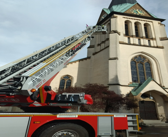 Feuerwehrfahrzeug vor einer Kirche mit einem Baum im Vordergrund und einem Kirchturm im Hintergrund unter einem bewölkten Himmel.