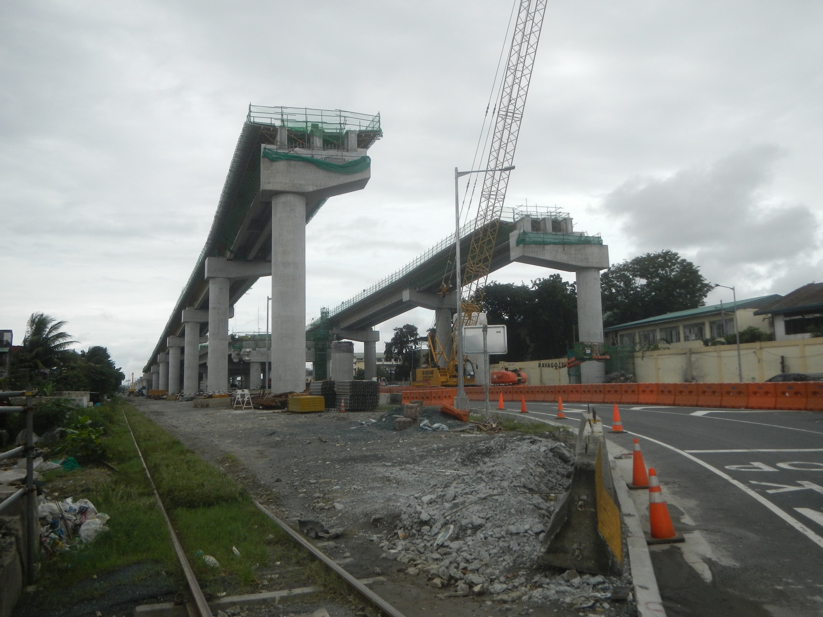 Baustelle mit einer Brücke im Hintergrund, eine Straße mit Absperrbaken auf der rechten Seite, Steine und Gras am Boden, eine Eisenbahnschiene auf der linken Seite, Bäume und Gebäude auf beiden Seiten und ein bewölkter Himmel.