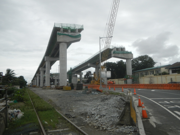 Baustelle mit einer Brücke im Hintergrund, eine Straße mit Absperrbaken auf der rechten Seite, Steine und Gras am Boden, eine Eisenbahnschiene auf der linken Seite, Bäume und Gebäude auf beiden Seiten und ein bewölkter Himmel.
