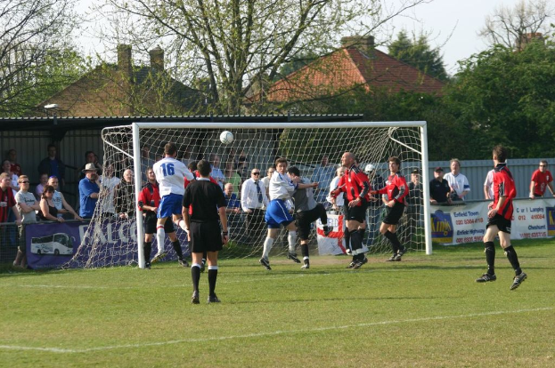 Fußballspieler spielen auf einem Feld mit einem Tornetz, während Zuschauer dahinter stehen, mit Bäumen und Häusern im Hintergrund.