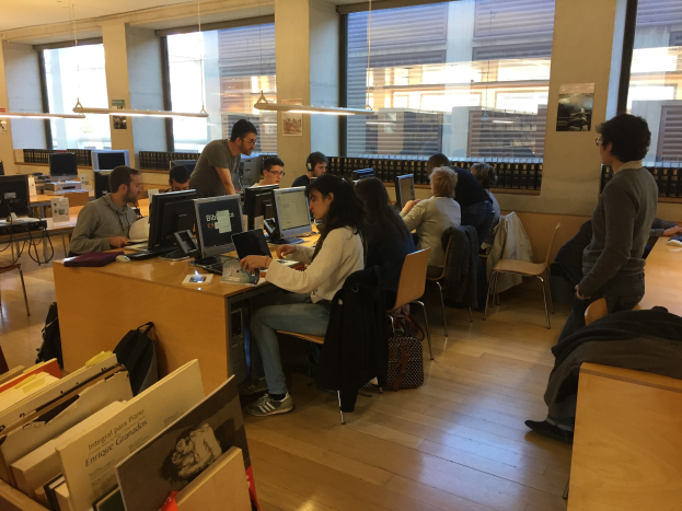 Eine Gruppe von Menschen, die an Computern auf Schreibtischen in einer Bibliothek arbeiten, die von Bücherregalen umgeben ist, mit Monitoren, Tastaturen und Taschen, unter Deckenlampen und in der Nähe von Glasfenstern.