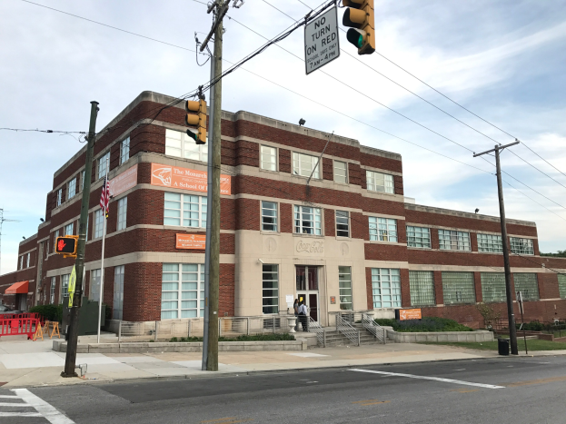 Großes Backsteingebäude mit der Aufschrift "Monarchs A-School" an einer Straßenkreuzung, mit Treppen, Geländern, Schildern und einer Person auf dem Bürgersteig, umgeben von Bäumen und Strommasten unter einem bewölkten Himmel.