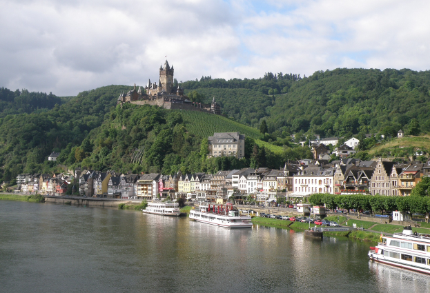 Ein idyllischer Blick auf den Rhein in Deutschland, mit einer Burg auf einem Hügel, Booten auf dem Fluss, Fahrzeugen auf einer näheren Straße und einem bewölktem Himmel.
