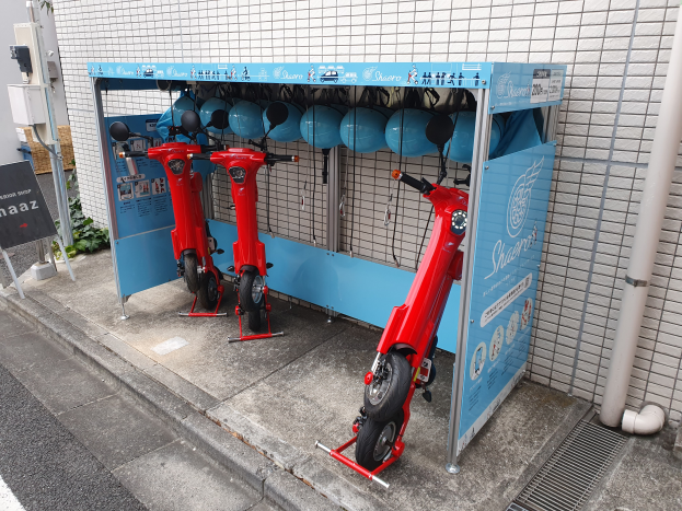Eine Reihe roter Roller auf einer Straße mit einem Stand, Rohren und einer Wand rechts daneben, Pflanzen und ein Schild im Hintergrund.