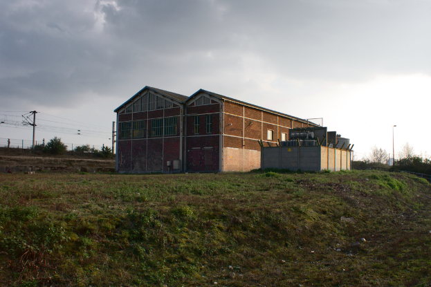 Eine Szene mit Hütten, Transformatoren, Strommasten, Stromkabel, Zäunen, einer Straßenlaterne, Straßenlaterne, Bäumen und einem bewölkten Himmel.