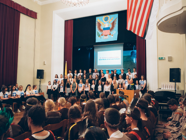 Eine Gruppe von Menschen steht vor einem sitzenden Publikum an einem Podium mit einem Mikrofon, flankiert von Fahnen und Lautsprechern, während eines 2. Wochen Kunst- und Kultur-Events.