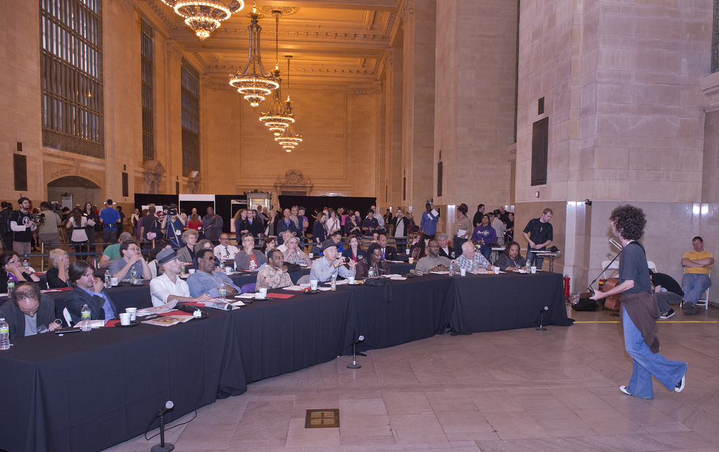 Ein Konzertbild mit einer Menge, einem Mann, der rechts Gitarre spielt und singt, einer Wand und Fenstern im Hintergrund, Lichtern oben und einem Boden unten.