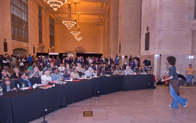 Ein Konzertbild mit einer Menge, einem Mann, der rechts Gitarre spielt und singt, einer Wand und Fenstern im Hintergrund, Lichtern oben und einem Boden unten.