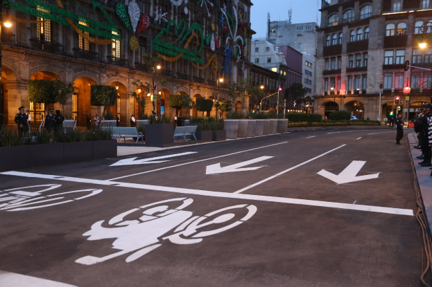 Stadtstraße mit einem ausgewiesenen Fahrradweg, umgeben von Gebäuden und Straßenmöbeln, Menschen auf dem Gehweg und einem klaren blauen Himmel.